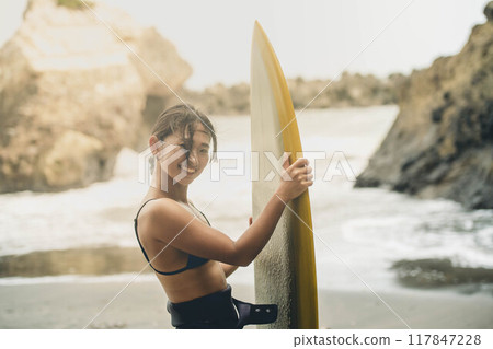 在風大的大海中拿著衝浪板站立的年輕女子 在風大的大海中拿著衝浪板站立的年輕女子 117847228