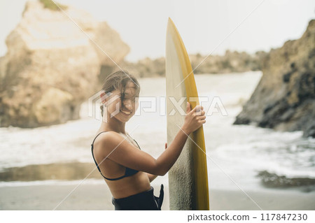 Young woman standing with surfboard in windy ocean Young woman standing with surfboard in windy ocean 117847230