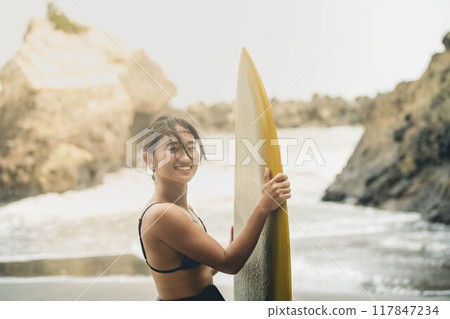 在風大的大海中拿著衝浪板站立的年輕女子 在風大的大海中拿著衝浪板站立的年輕女子 117847234