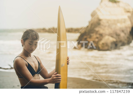 在風大的大海中拿著衝浪板站立的年輕女子 在風大的大海中拿著衝浪板站立的年輕女子 117847239