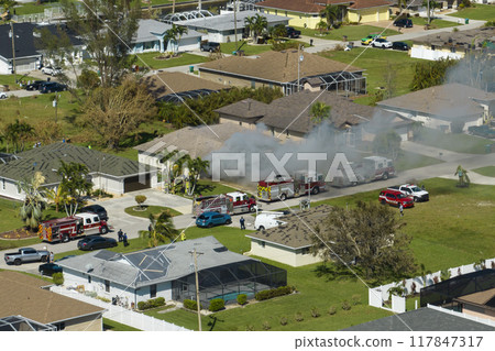 Aerial view of house on fire and firefighters extinguishing flames after short circuit caused spark to ignite wooden roof damaged by hurricane Ian wind. Home disaster in Florida suburban area Aerial view of house on fire and firefighters extinguishing flames after short circuit caused spark to ignite wooden roof damaged by hurricane Ian wind. Home disaster in Florida suburban area 117847317