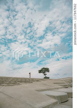 A young woman walking next to a tree on the beach under a blue sky A young woman walking next to a tree on the beach under a blue sky 117847682