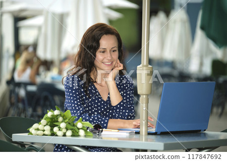 Young happy woman working remotely on laptop computer sitting at outdoor restaurant table. Doing business online concept Young happy woman working remotely on laptop computer sitting at outdoor restaurant table. Doing business online concept 117847692