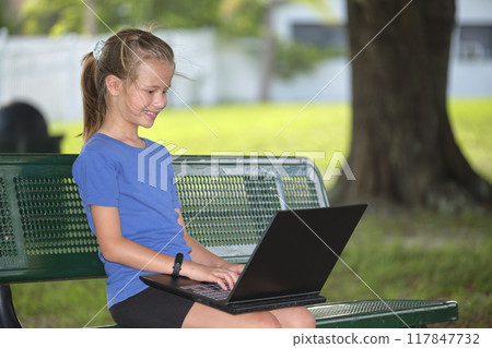 Schoolgirl studying on laptop computer resting on park bench outdoors on summer day. Remote education during quarantine concept 117847732