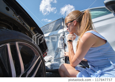 Sad young woman driver sitting near her smashed car looking shocked on crashed vehicles in road accident 117847737