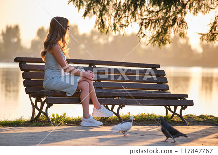 Lonely woman sitting on lake side bench enjoying warm summer evening. Solitude and relaxation concept. Lonely woman sitting on lake side bench enjoying warm summer evening. Solitude and relaxation concept. 117847767