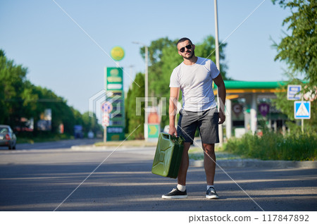Confident man finally found petrol station and fill canister. Man returning to car to refuel it. Young male with canister in hand on background of gas station. 117847892