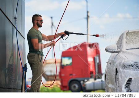 Washing luxury white auto with soap gun on an open air car wash. Confident man cleaning his car with foam. Technical service concept. 117847893