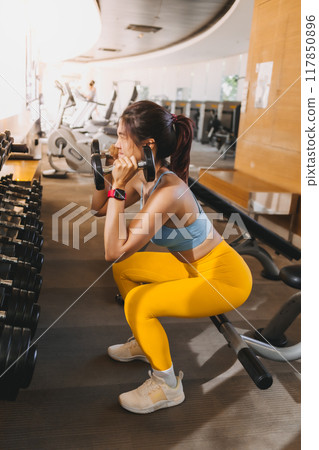 Woman wears blue and yellow outfits do dumbbell squats in the gym. 117850896