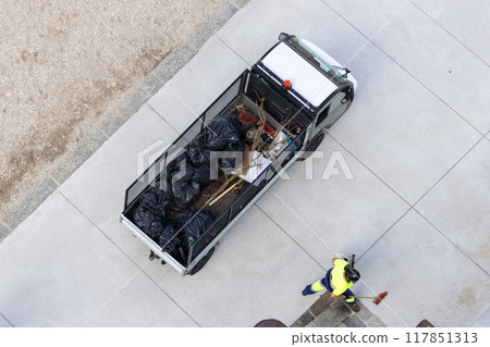 Aerial view of a Electric vehicle loaded with garbage bags and a street cleaner working to collect trash in a city beach area 117851313
