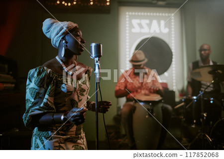 Young Black woman wearing stylish dress with headwrap singing in microphone during performance with jazz band 117852068