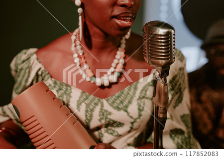 Medium closeup of young African American woman holding guiro in hands singing jazz song in microphone 117852083