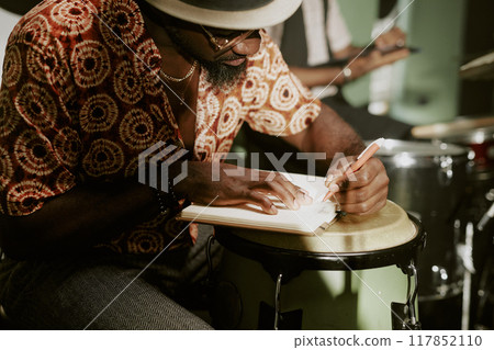 African American musician sitting at tumbadora writing note in notebook during rehearsal with jazz band African American musician sitting at tumbadora writing note in notebook during rehearsal with jazz band 117852110