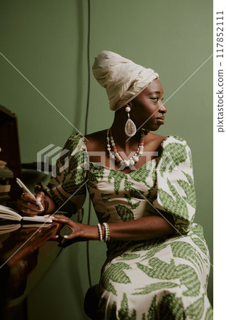 Vertical medium long shot of young African American female musician sitting at piano looking away while writing lyrics for new song Vertical medium long shot of young African American female musician sitting at piano looking away while writing lyrics for new song 117852111