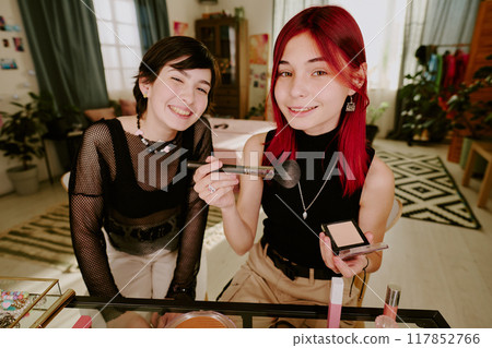 Two friends happily sharing makeup tips while sitting in cozy and well-decorated living room surrounded by mirrors, brushes, and cosmetics, creating a joyful and friendly atmosphere Two friends happily sharing makeup tips while sitting in cozy and well-decorated living room surrounded by mirrors, brushes, and cosmetics, creating a joyful and friendly atmosphere 117852766