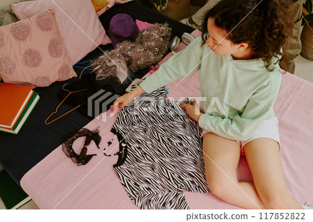 Young girl with long curly hair selecting zebra-print shirt on bed filled with other clothes and accessories Young girl with long curly hair selecting zebra-print shirt on bed filled with other clothes and accessories 117852822