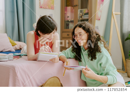 Two teenagers studying together in cozy bedroom, one making notes while other holds book and laughs. Warm, inviting ambiance with pastel decoration and desk in background 117852854