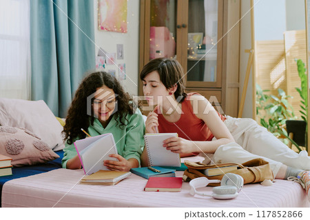 Two friends sitting on a comfortable bed, engaged in studying with notebooks, pens, and other study materials spread around them Two friends sitting on a comfortable bed, engaged in studying with notebooks, pens, and other study materials spread around them 117852866