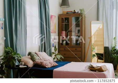 Cozy bedroom featuring houseplants and a bookshelf filled with books and decorative items, bathed in sunlight streaming through windows with blue curtains Cozy bedroom featuring houseplants and a bookshelf filled with books and decorative items, bathed in sunlight streaming through windows with blue curtains 117852875