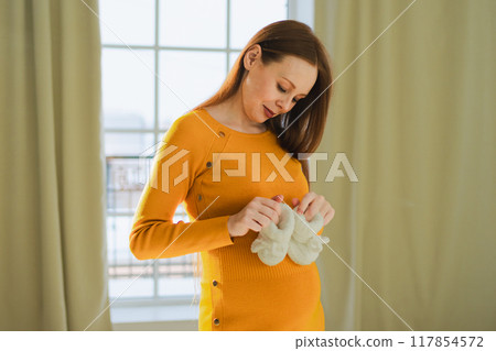 Pregnancy motherhood people expectation future. Pregnant woman with big belly holding newborn baby booties smiling at home. Young mom enjoying pregnancy. Maternity tenderness parenthood new life Pregnancy motherhood people expectation future. Pregnant woman with big belly holding newborn baby booties smiling at home. Young mom enjoying pregnancy. Maternity tenderness parenthood new life 117854572