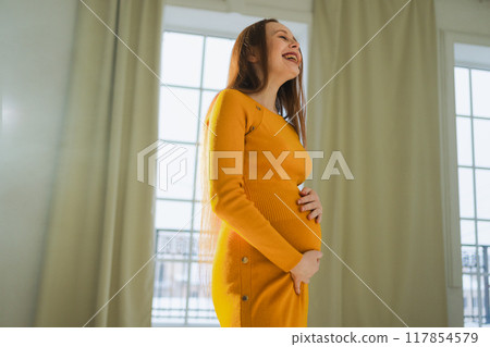 Pregnancy motherhood people expectation future. Pregnant woman with big belly standing near window at home. Girl hugging her tummy enjoying pregnancy. Maternity tenderness parenthood new life concept 117854579