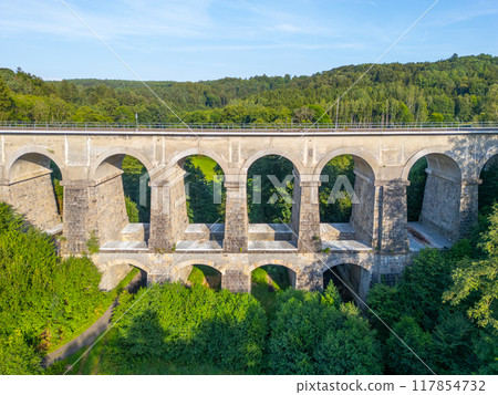 The Sychrov Railway Bridge rises gracefully above lush greenery, showcasing its impressive arches against a backdrop of vibrant trees. 117854732