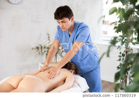 Professional young masseur performing back massage for female patient Professional young masseur performing back massage for female patient 117860319