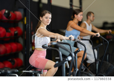 Girl engaged in air bike workout with group of sporty people in exercise room during weight training classes 117860338