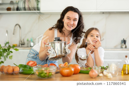 Mother holding saucepan and preteen daughter standing at table 117860341