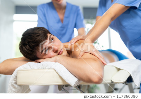 Young man relaxing while two masseuses doing four hands back massage Young man relaxing while two masseuses doing four hands back massage 117860380