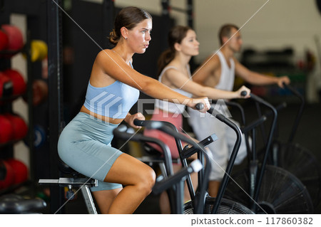 Girl engaged in air bike workout with group of sporty people in exercise room during weight training classes Girl engaged in air bike workout with group of sporty people in exercise room during weight training classes 117860382