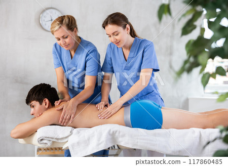 Two female masseuses perform stroking and rubbing of muscles of back and shoulders to guy client 117860485