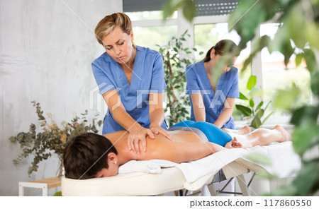Masseuse with female assistant performing four hands massage to man 117860550