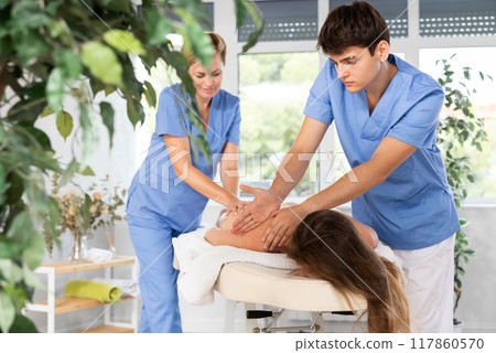 Young masseur in tandem with female colleague performing back massage to woman Young masseur in tandem with female colleague performing back massage to woman 117860570