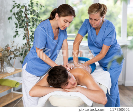 Young masseuse trainee with experienced female mentor doing back massage to man 117860601