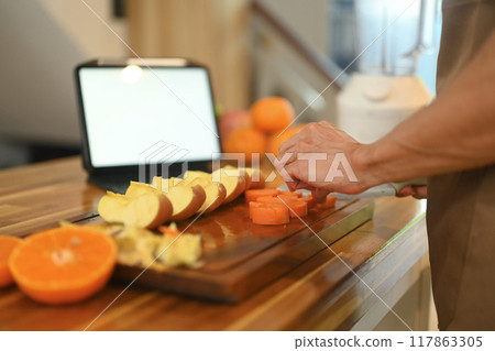 Man cutting up fruit on a wooden cutting board and looking recipes on digital tablet Man cutting up fruit on a wooden cutting board and looking recipes on digital tablet 117863305