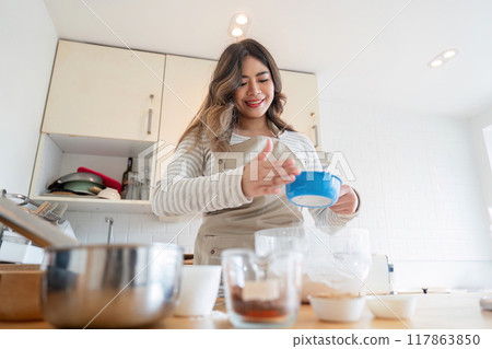Smiling Asian woman adding ingredients while baking in kitchen 117863850