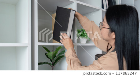 Asian housekeeper organizing books on office shelf with care 117865247