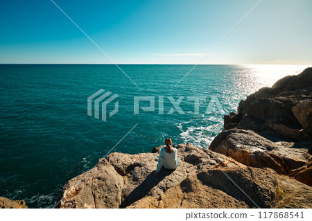 A girl sits on the edge of a cliff and looks at the sea A girl sits on the edge of a cliff and looks at the sea 117868541