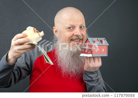 bald man with a long gray beard holding a piggy bank with a dollar bill and a small model house bald man with a long gray beard holding a piggy bank with a dollar bill and a small model house 117870088