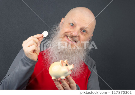 mature man with a long gray beard smiling while putting a coin into a piggy bank 117870301