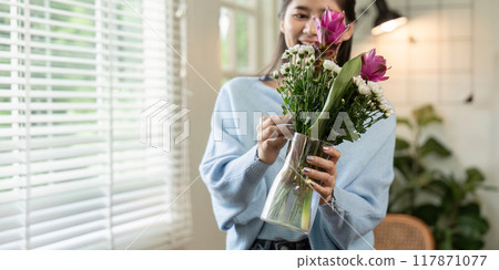 Cheerful Asian woman arranging fresh flowers in sunlit home Cheerful Asian woman arranging fresh flowers in sunlit home 117871077