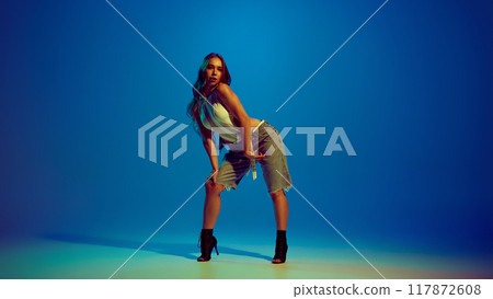 Sensual young beautiful woman dancing high heel dance, performing against blue studio background in neon light Sensual young beautiful woman dancing high heel dance, performing against blue studio background in neon light 117872608