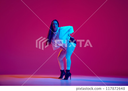 Sensual young woman in oversized t shirt and black heeled shoes dancing high heel dance against magenta studio background 117872648