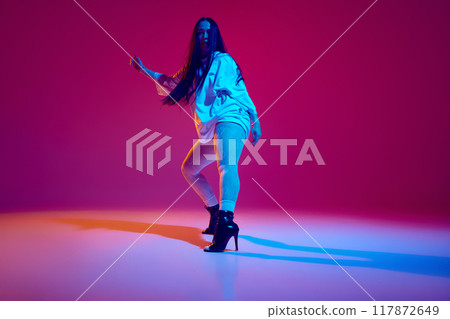 Artistic, talented, beautiful young woman in oversized clothes and heeled shoes dancing high heel dance against magenta studio background 117872649