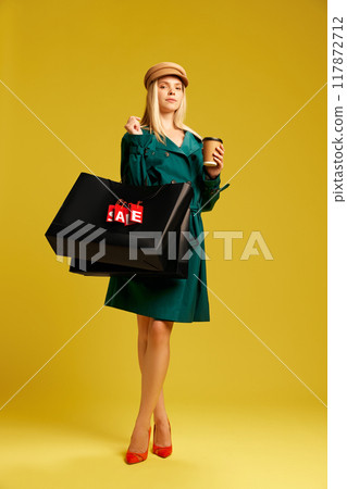 Elegant young woman in green trench coat carrying black shopping bags and drinking coffee against yellow studio background 117872712