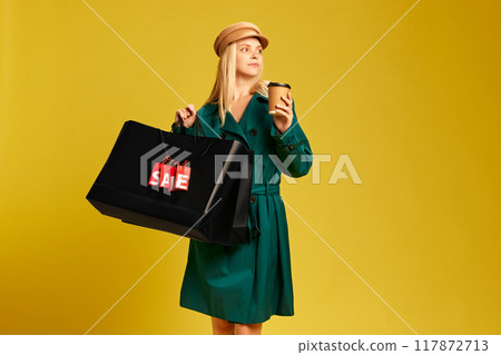 Elegant young woman in green trench coat carrying black shopping bags and drinking coffee against yellow studio background 117872713