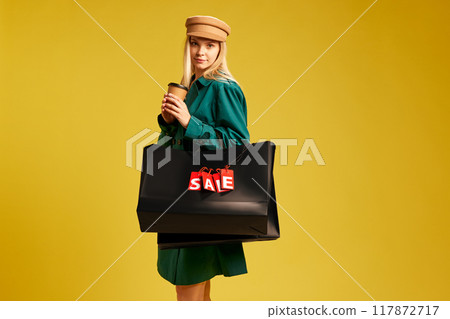 Beautiful young woman in stylish green coat standing with black shopping bags against yellow studio background. Autumn sales 117872717