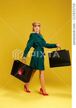 Elegant young woman in green coat cheerfully carrying black shopping bags after successful shopping against yellow studio background 117872719
