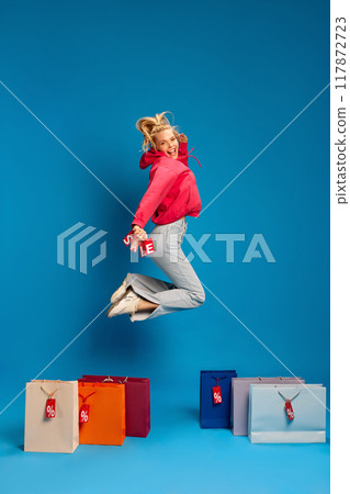 Happy young woman in casual clothes jumping around many colorful shopping bags with percentage signs on it against blue studio background 117872723